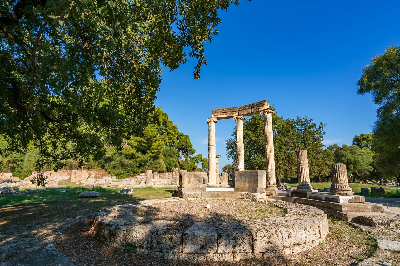Ancient stone columns and ruins of the Temple of Zeus at Olympia, surrounded by trees under a clear blue sky.